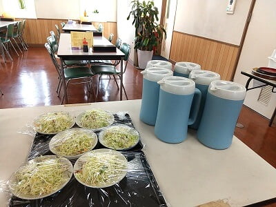 港湾食堂 和以尊の横のテーブルにお冷のポットに1人分ずつお皿に乗ってラップがかかってるサラダがある