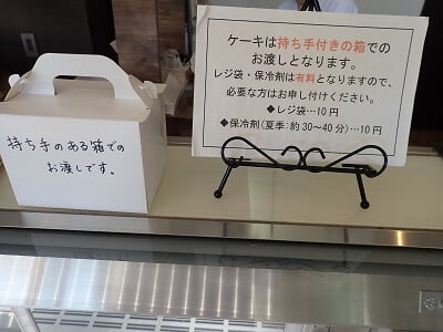 Patisserie HIMITSUKICHI(ヒミツキチ)の正面のケーキのショーケース上に「ケーキは持ち手のある箱でお渡し」でレジ袋、保冷剤は有料と案内